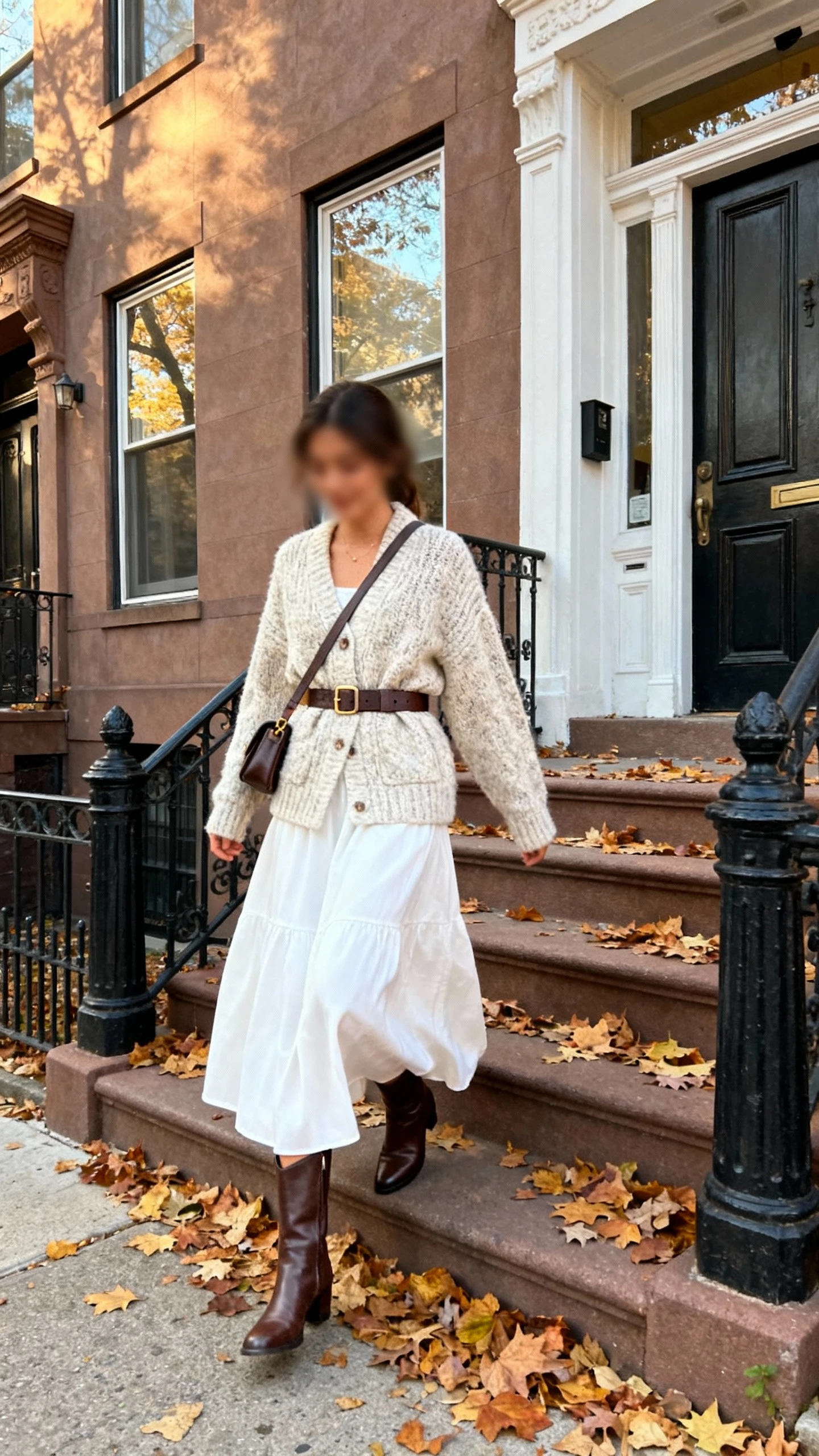 Candid photo of a woman wearing a white maxi skirt with a chunky cardigan cinched by a belt, mid-calf boots, and a crossbody bag, stepping onto a townhouse stoop with fallen leaves, face slightly blurred, soft afternoon light, iPhone photo quality.