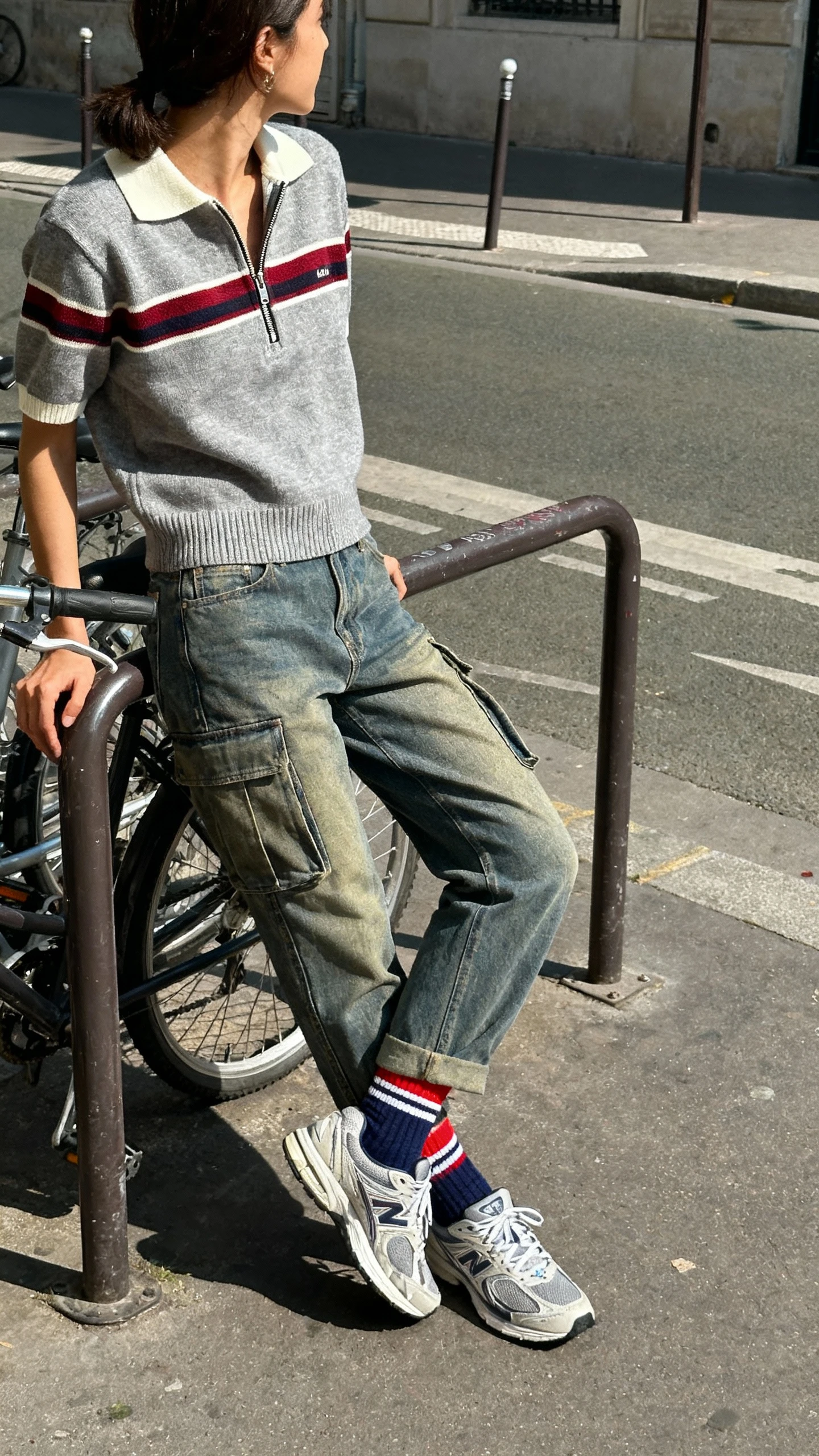 Natural lifestyle photo of a woman in cargo jeans, retro zip polo knit, retro running sneakers, and sporty tube socks, leaning against a bike rack, face turned away, sunny afternoon light, iPhone candid.