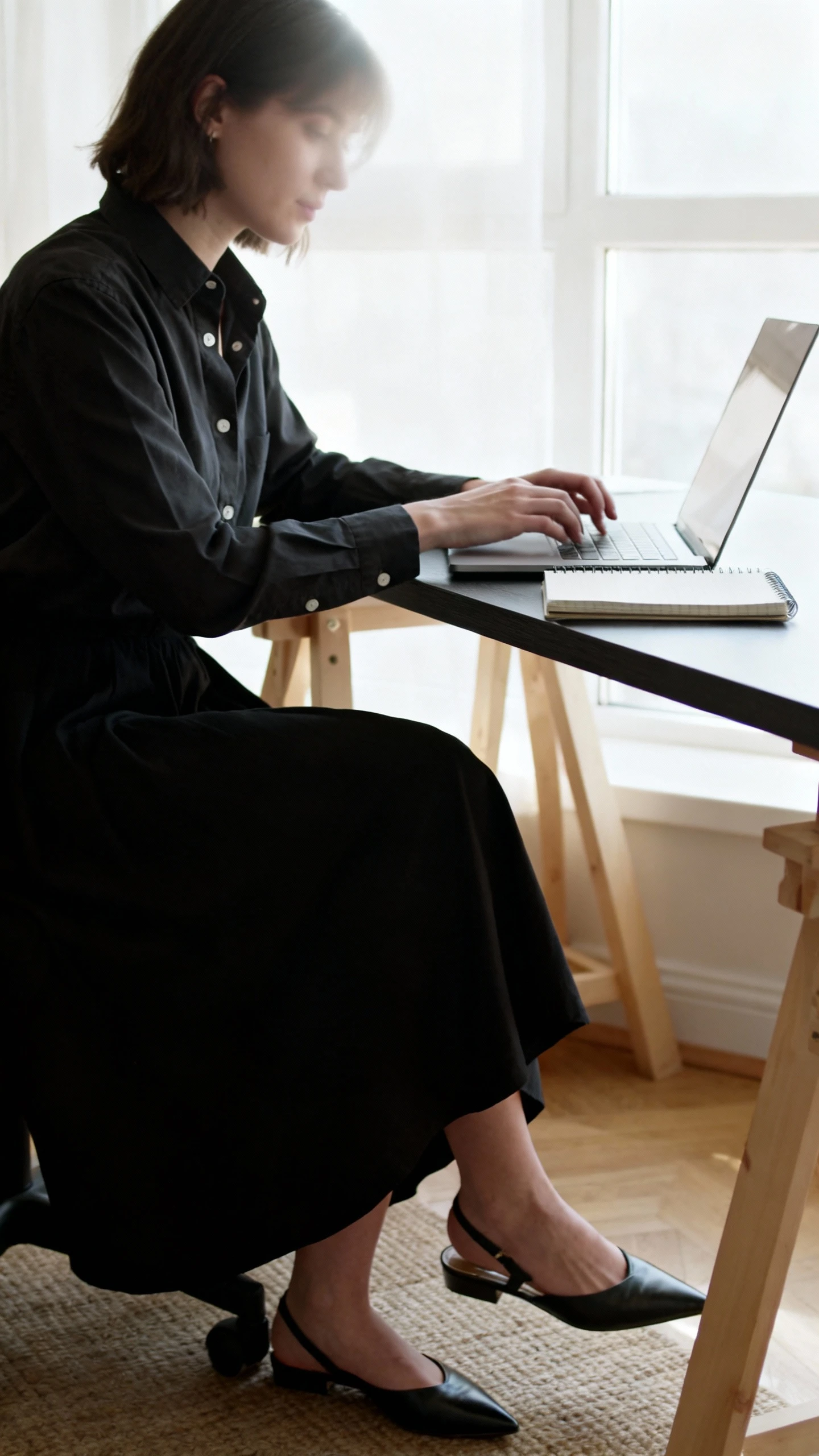 Office candid of a woman in a black maxi skirt with a crisp button-up shirt and pointed flats, working at a desk with a laptop and notebook, face slightly blurred, soft window light, iPhone photo look.