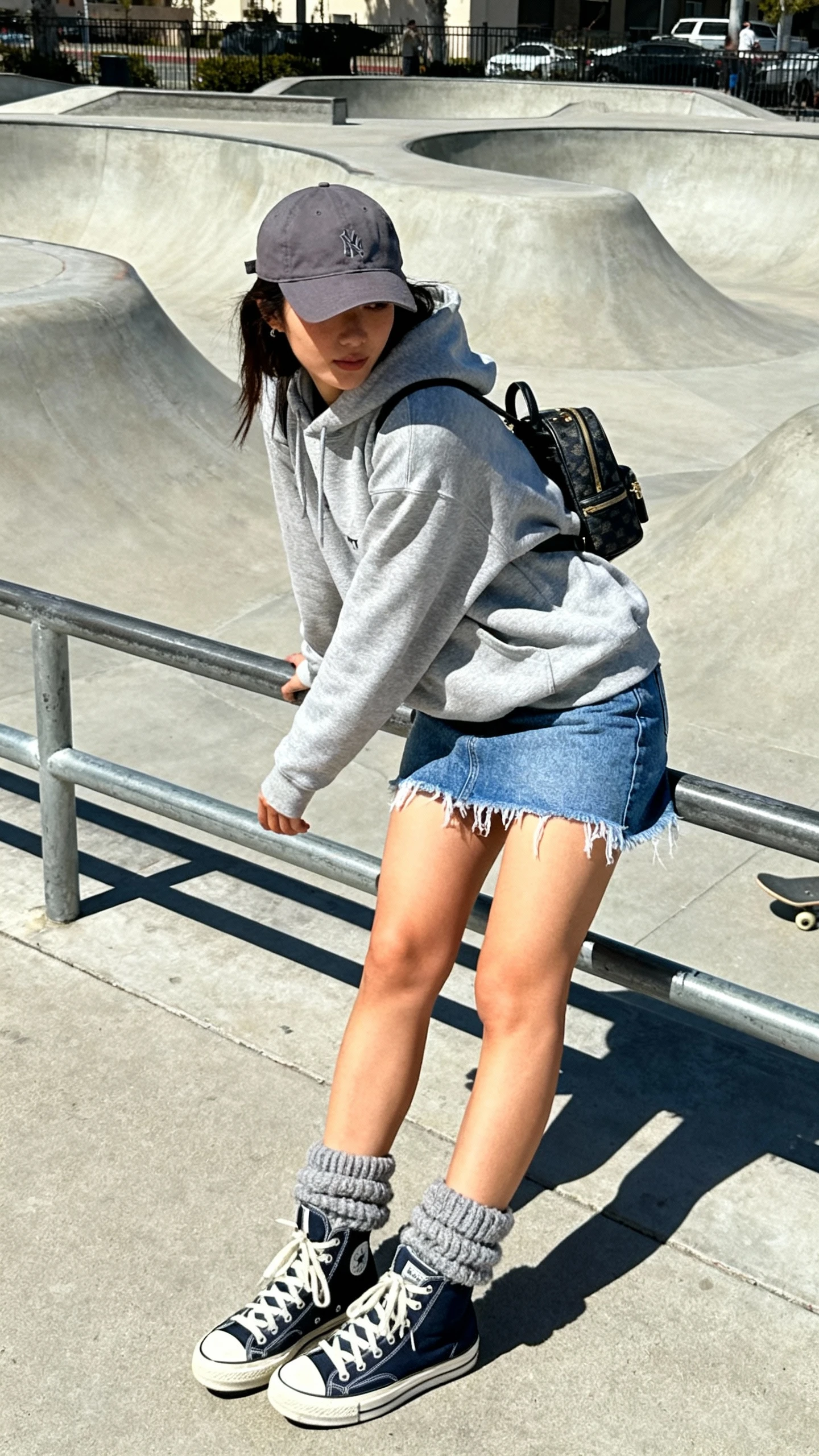 Urban candid of a woman in a gray hoodie, raw-hem denim mini skirt, high-top sneakers, baseball cap, mini backpack, chunky socks peeking out, leaning on a railing by a skatepark, face in shadow, afternoon daylight, iPhone photo quality.