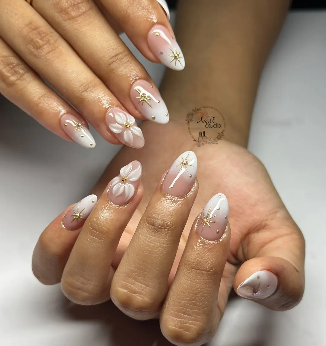 Nude-and-white almond nails with gold starburst charms and raised white flowers