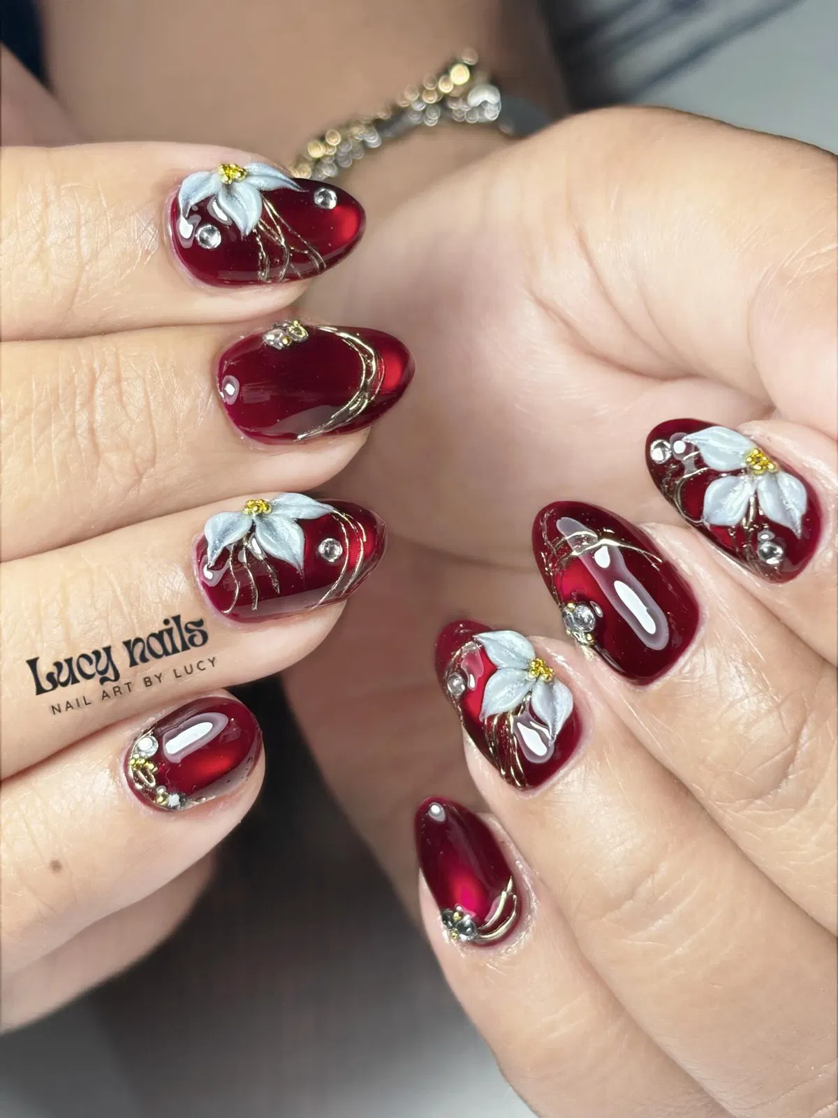 Deep red rounded nails with white flowers, gold lines, and rhinestones