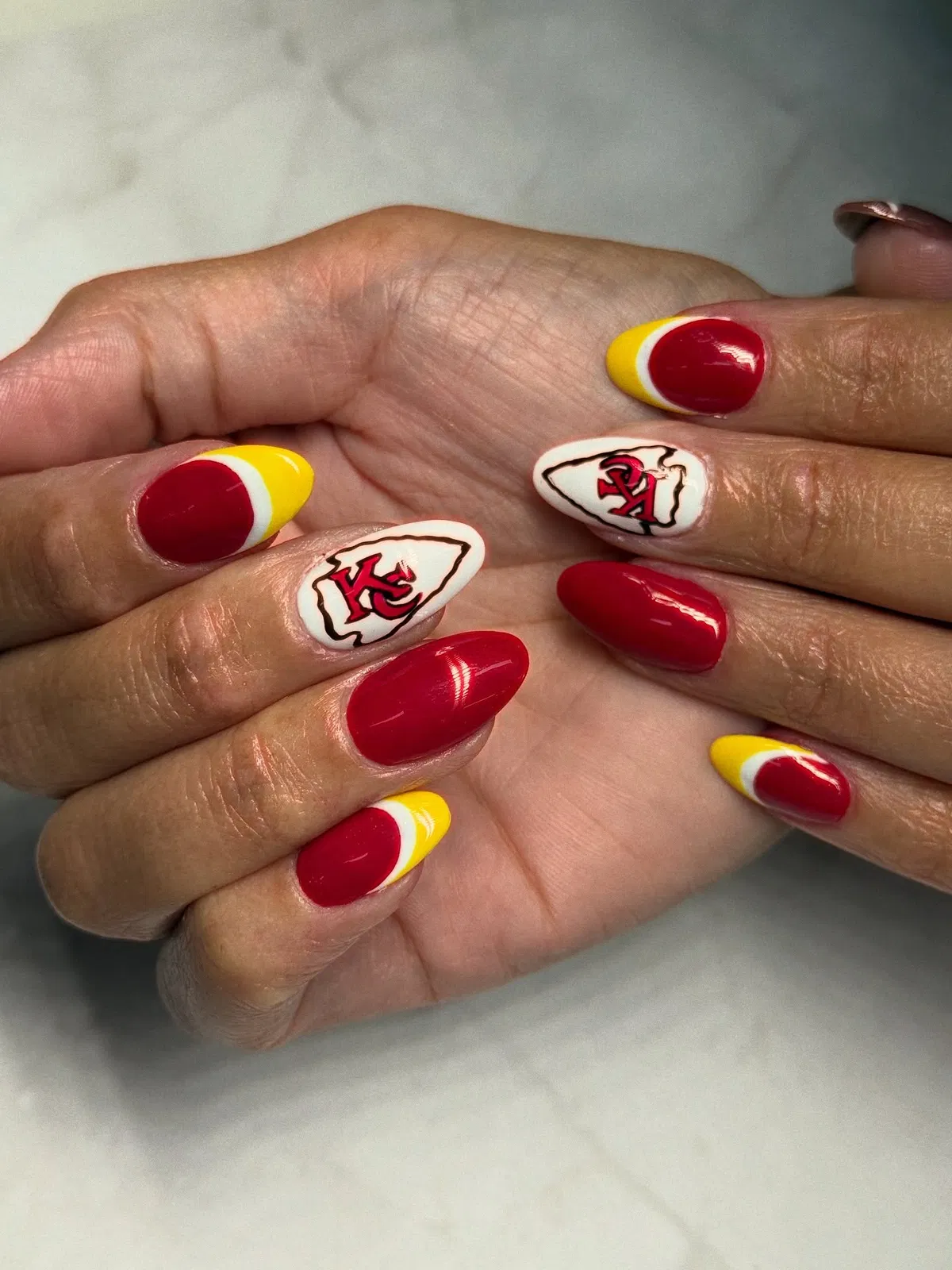 Red almond nails with 'KC' arrowhead accents and yellow-tipped details