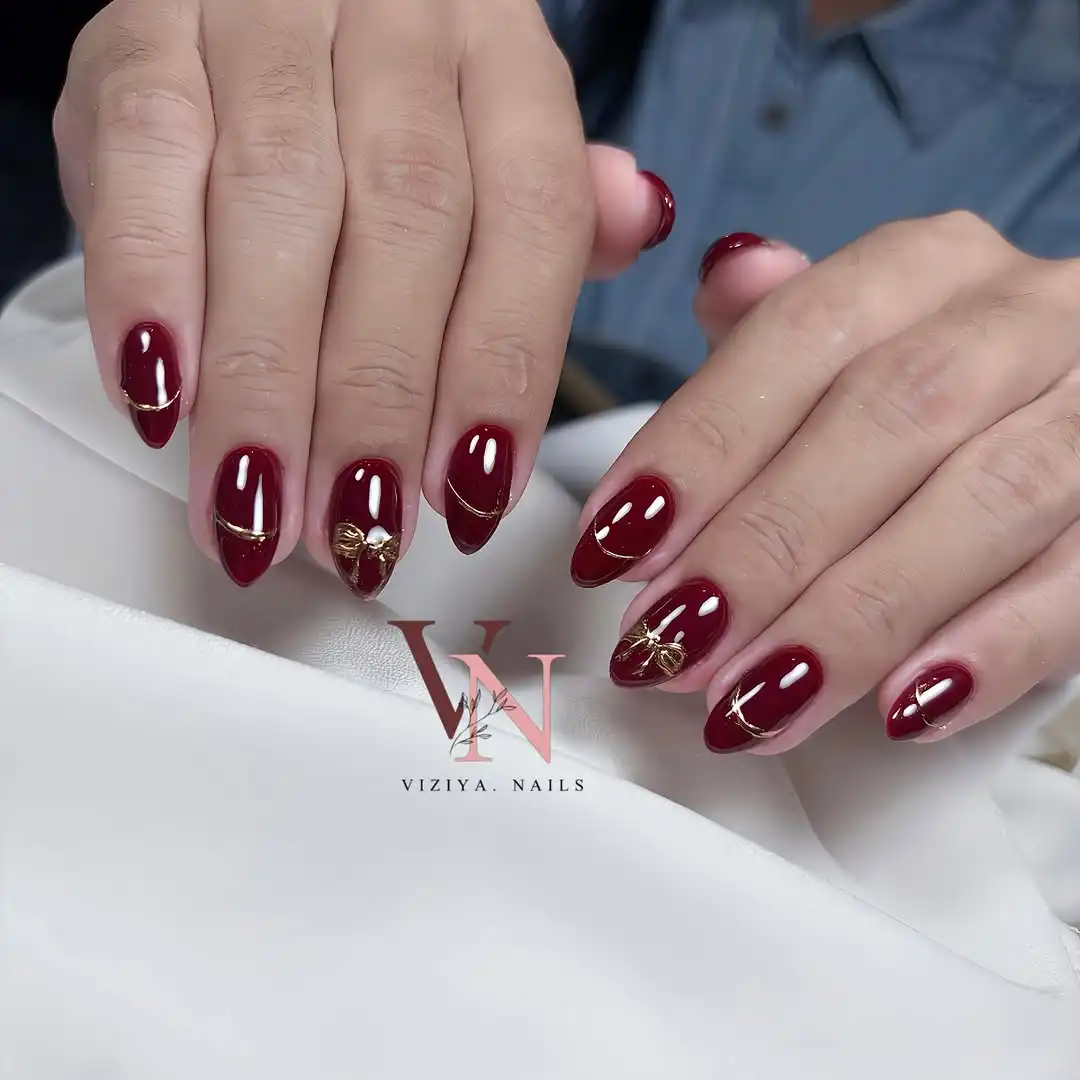 Maroon nails with curved gold lines and bow accents