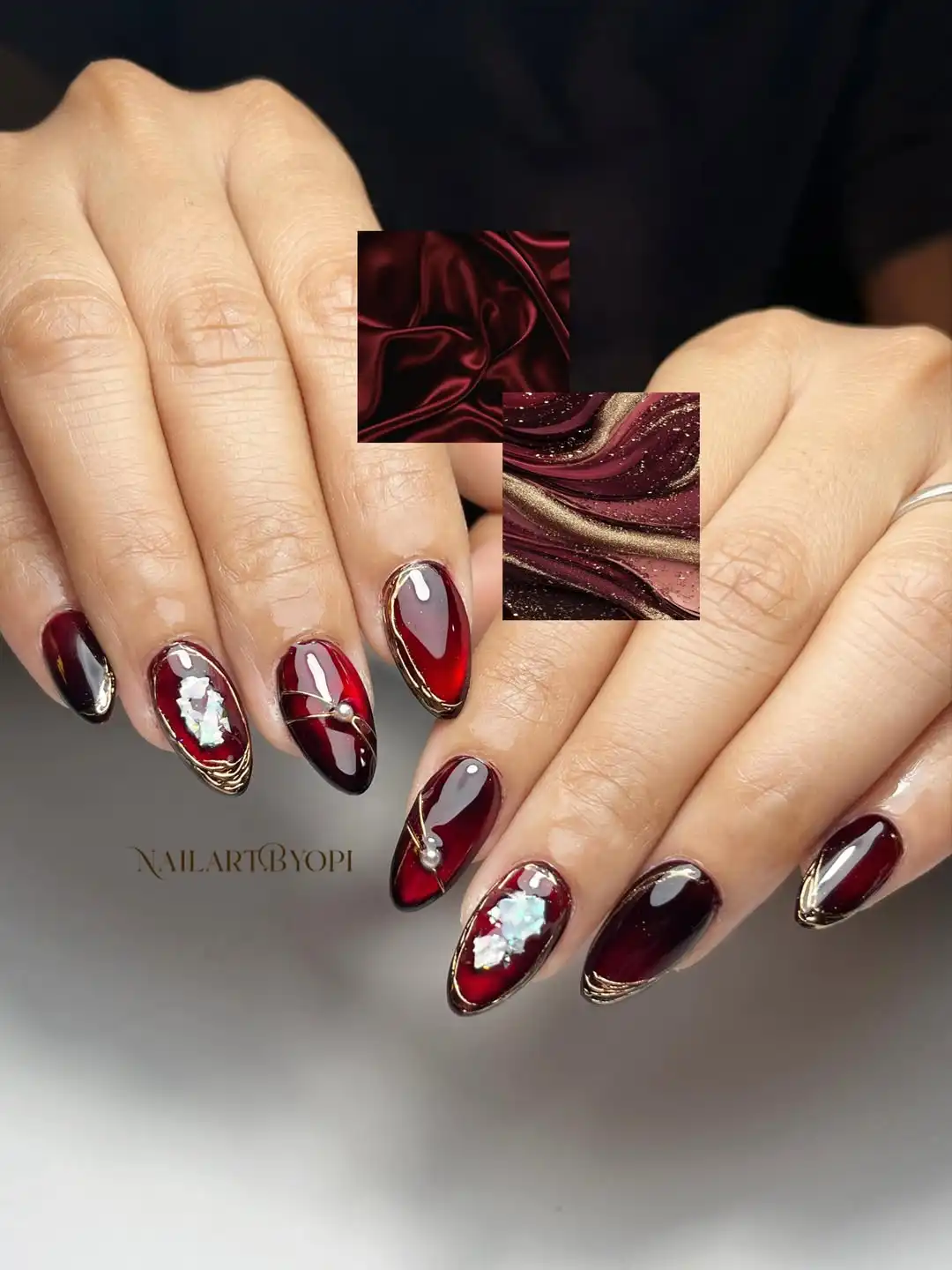 Deep maroon almond nails with gold edging, reflective accents, and a pearl-like detail