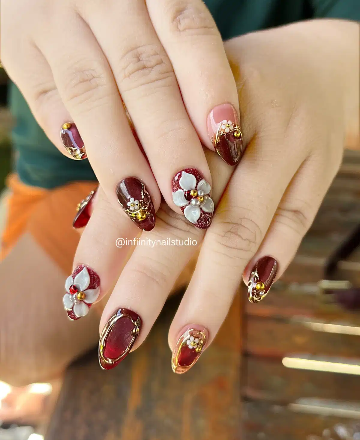 Deep maroon nails with gold framing and raised white floral accents