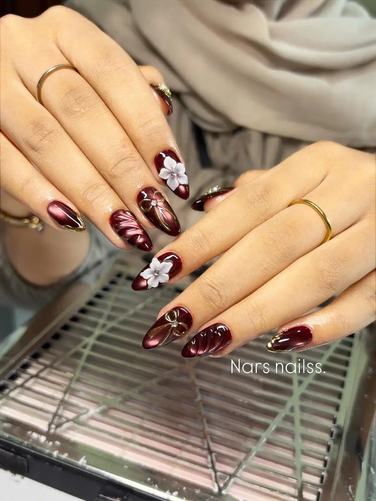 Reflective maroon nails with gold bow lines and 3D white flowers