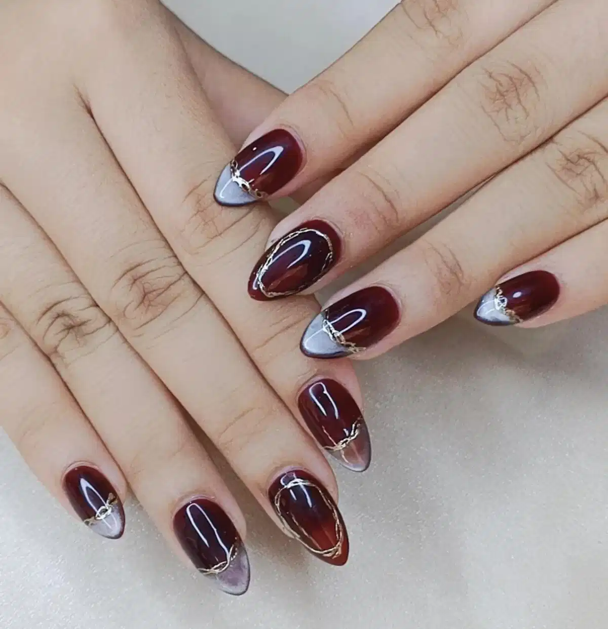 Maroon almond nails with silver-gray triangle tips and thin gold outlines